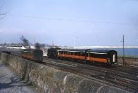 A smokey Crossley engined C Class locomotive No.C212 is seen shunting some parcal vans at the north end of Dun Laoghaire Station. At this time in the early 1970s Victorian coaching stock was still used on services in the Dublin area, such as the ex Great Southern &#38; Western carriage stabled in the siding. The level crossing gates for the Caol Quay siding can also be seen in front of the loco. ©Barry Carse
